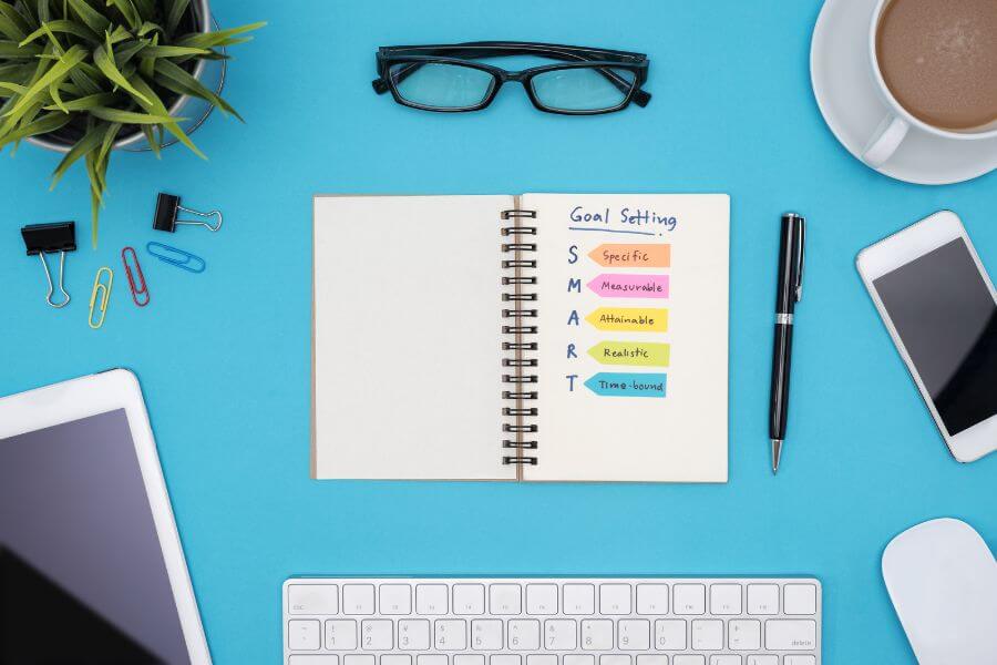 SMART Goal Setting for Social Media - Light blue desktop with plants, keyboard, glasses coffee cup, devices and notepad outlining SMART.